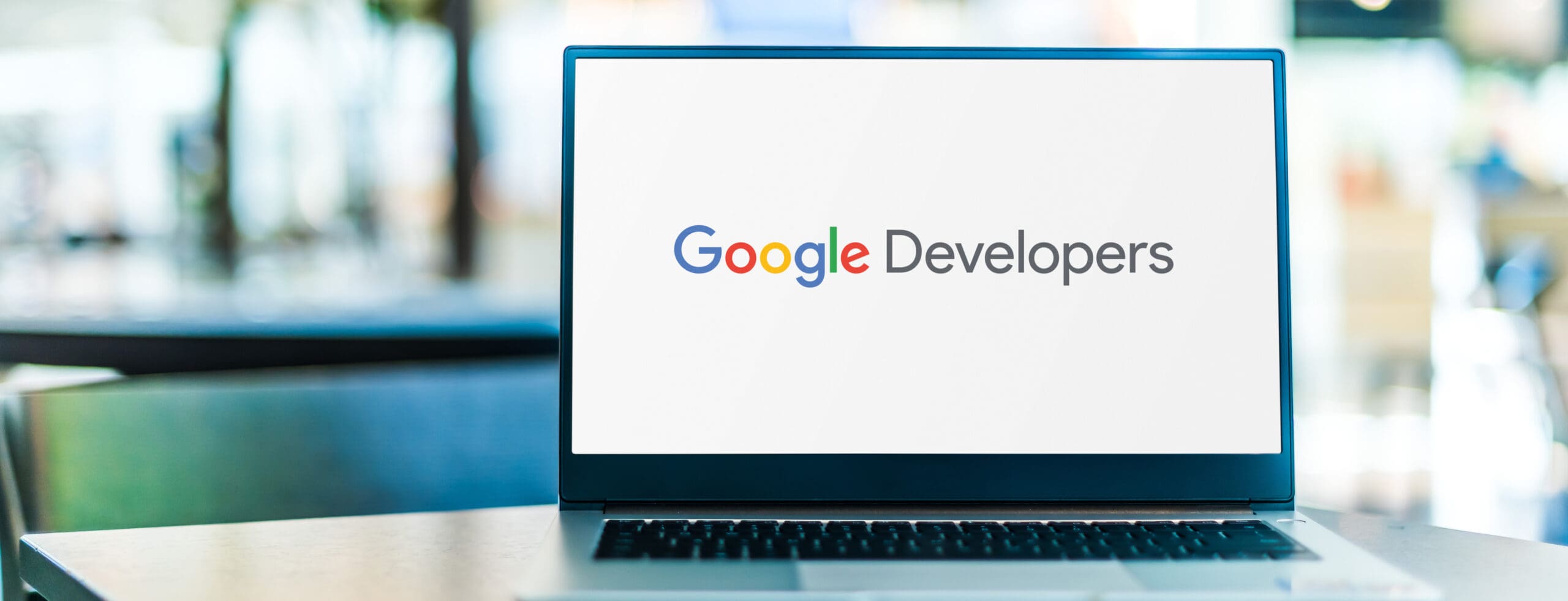 A laptop screen displays the Google Developers logo, with the words "Google Developers" in colorful text. The background is blurred, suggesting a modern indoor setting.